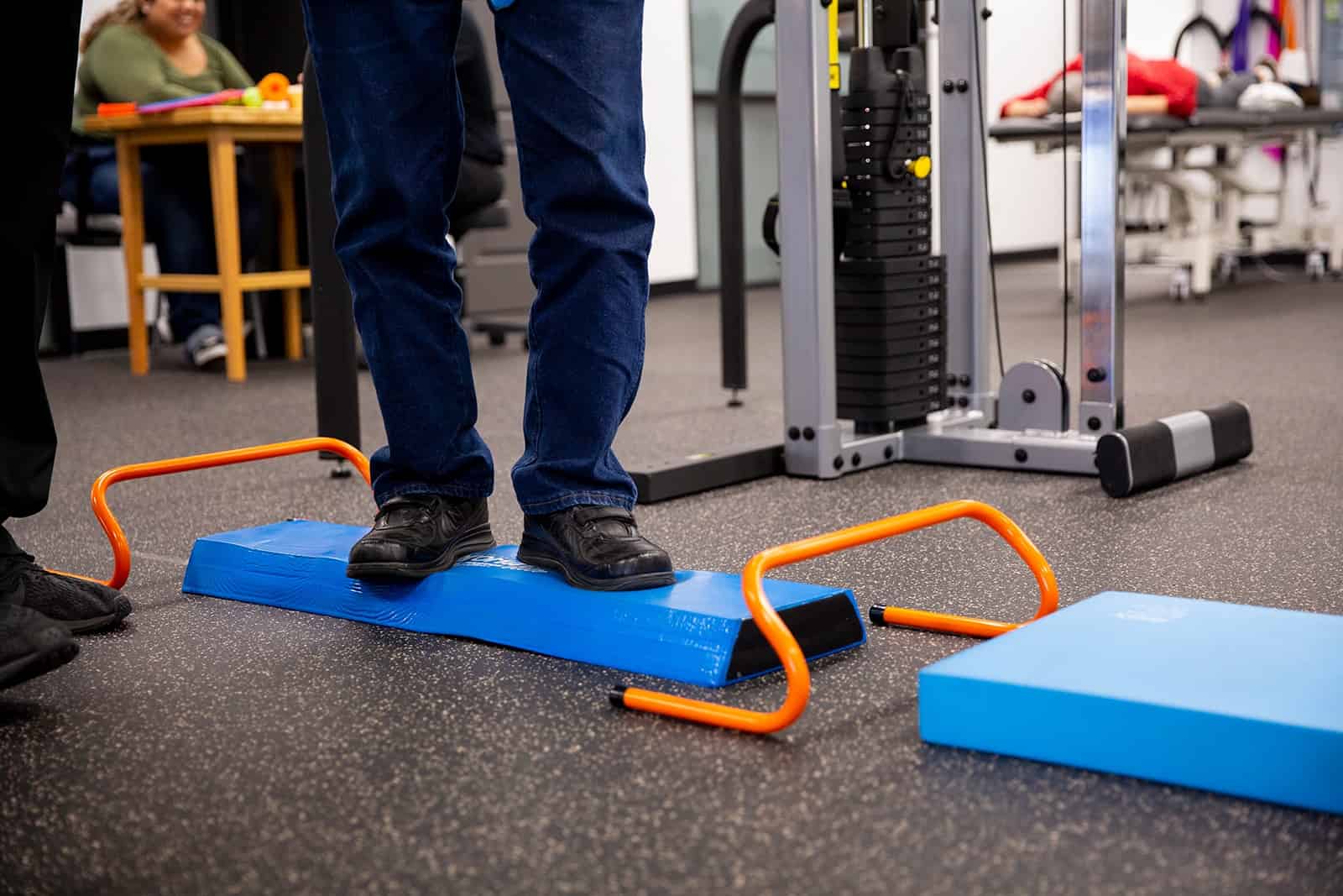 Person practicing balance exercises on a blue balance pad with orange handles in a gym setting.