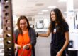 A female physical therapy assistant at PRO~PT Fresno West in black clothes smiles as she helps a female patient in an orange shirt and cardigan during an exercise with flexible bands.