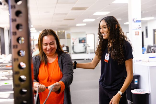A female physical therapy assistant at PRO~PT Fresno West in black clothes smiles as she helps a female patient in an orange shirt and cardigan during an exercise with flexible bands.