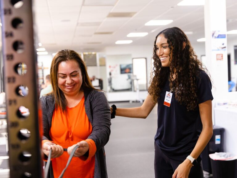 A female physical therapy assistant at PRO~PT Fresno West in black clothes smiles as she helps a female patient in an orange shirt and cardigan during an exercise with flexible bands.